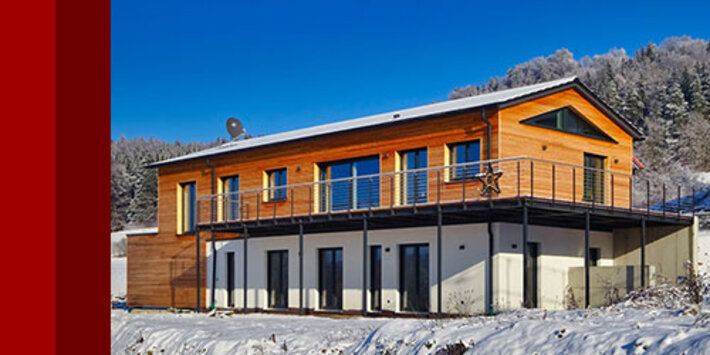 Haus WM im Winter: Holzfassade mit umlaufendem Balkon vor verschneitem Hang