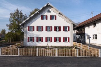 Giebelseite des Dorfgemeinschaftshauses mit roten Fensterläden und Gartenzaun