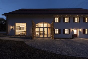 Dorfgemeinschaftshaus Gutenberg in der Dämmerung mit warm beleuchteten Fenstern und geöffnetem Holztor